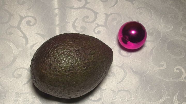Eine Reife Avocado liegt neben einer pinken Weihnachtskugel auf einem eleganten, gl&auml;nzenden Tischtuch.