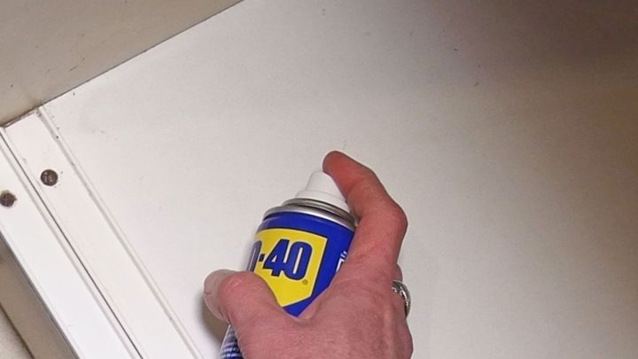 Eine Hand h&auml;lt eine Spraydose, um fettige K&uuml;chenregale zu reinigen, w&auml;hrend sie in einer hellen K&uuml;chenschrankfl&auml;che wirkt.
