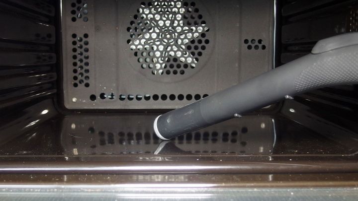 Ein Staubsaugeraufsatz entfernt Kr&uuml;mel im Inneren eines Ofens, w&auml;hrend der gl&auml;nzende Innenraum gut sichtbar ist.