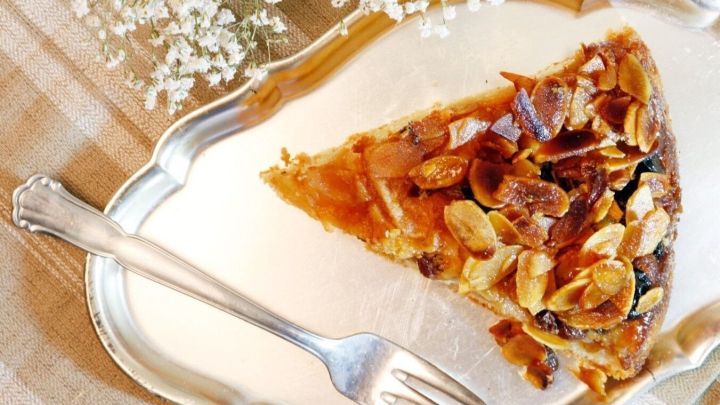 Ein St&uuml;ck Apfeltorte liegt auf einem silbernen Teller, w&auml;hrend eine Gabel daneben bereit ist, zu kosten. Im Hintergrund sind zarte Blumen und ein kariertes Tuch.