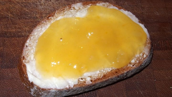 Ein St&uuml;ck Brot liegt auf einem Holzbrett, bestrichen mit leuchtend gelber Orangenmarmelade, die s&uuml;&szlig; und aromatisch aussieht.