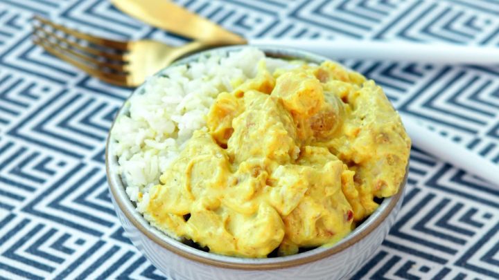 Eine Sch&uuml;ssel mit Curry-H&auml;hnchen in Pfirsich-Sahne liegt auf einem geometrischen Tischmuster, daneben Besteck.