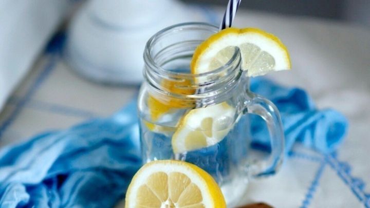 Ein Einmachglas mit Zitronenwasser und Strohhalm, umgeben von geschnittenen Zitronen auf einem Holzbrett, liegt auf einem blauen Tuch.