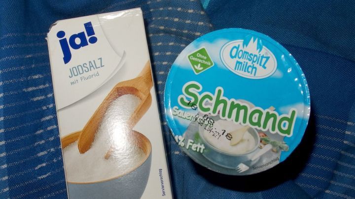 Schmandbecher und Jodsalzpackung liegen auf blauem Stoff; ideal f&uuml;r g&uuml;nstiges DIY-Bodypeeling zu Hause.