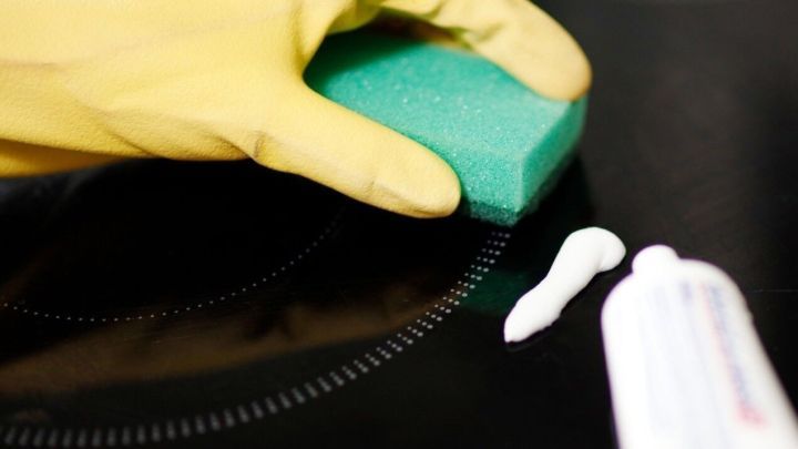 Ein gelber Handschuh h&auml;lt einen Schwamm, der auf einem Glaskeramikfeld mit Zahnpasta gereinigt wird.