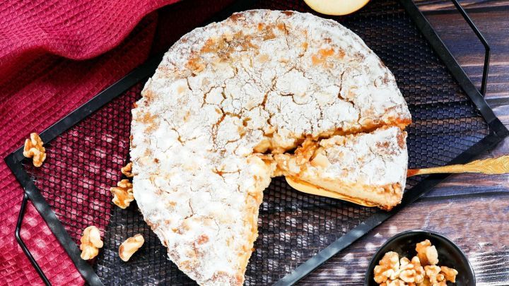 Ein Apfelkuchen mit Waln&uuml;ssen liegt auf einem gitterf&ouml;rmigen Tablett, w&auml;hrend ein St&uuml;ck herausgeschnitten ist.