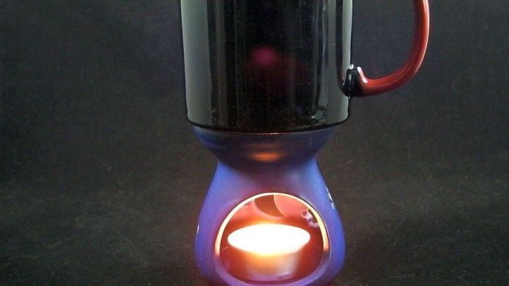 Ein Kaffeebecher steht auf einem Tassenw&auml;rmer ohne Strom, der durch eine Kerze beheizt wird, in dunklem Umfeld.