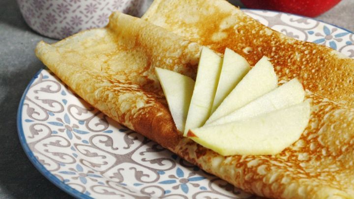 Eine goldene Pfannkuchenrunde liegt auf einem dekorativen Teller, belegt mit frisch geschnittenen Apfelscheiben, daneben ein roter Apfel.