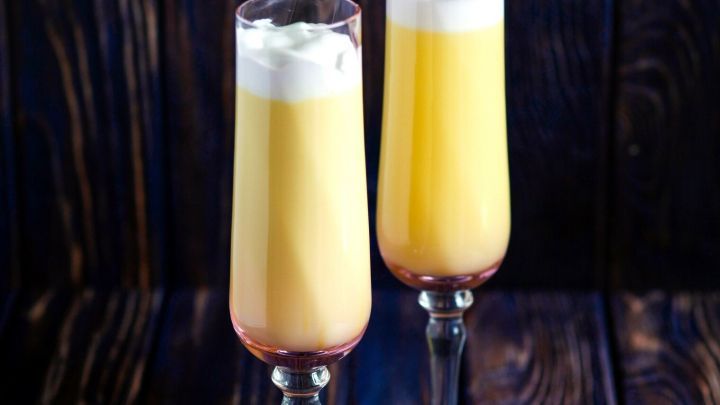 Zwei elegante Gl&auml;ser mit einem gelben Cocktail, geschichtet, auf einer dunklen Holzoberfl&auml;che, mit einer Sahnehaube dekoriert.