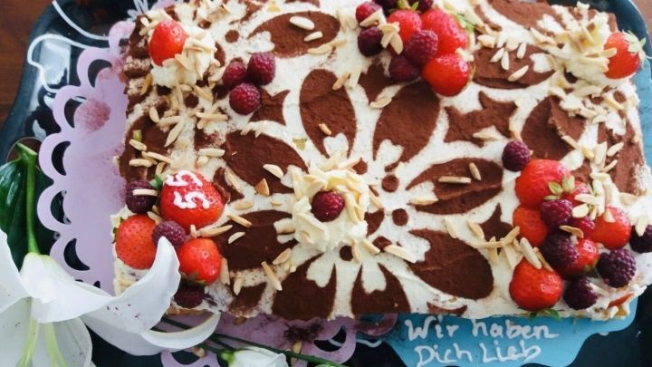 Ein kunstvoll verziertes St&uuml;ck Torte mit Mustern aus Sahne, frischen Beeren und Mandeln auf einem eleganten Teller.