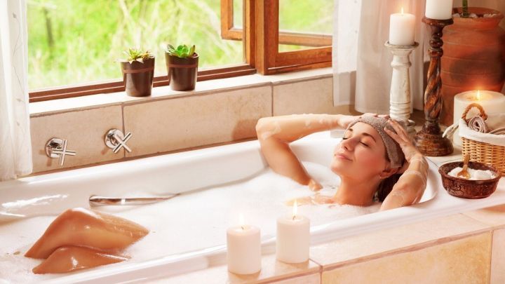 Eine Frau entspannt in einer Badewanne mit Wasser, umgeben von Kerzen und Pflanzen, w&auml;hrend sie Husten lindert.