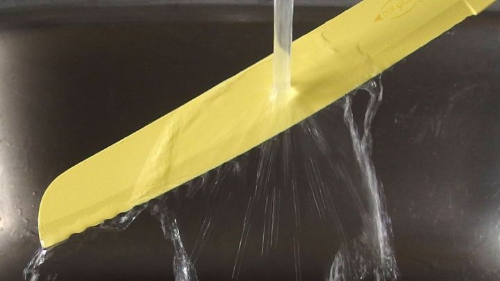 Ein gelbes Messer wird unter flie&szlig;endem Wasser gewaschen, was darauf hindeutet, dass es zum leichteren Schneiden von Brot vorbereitet wird.