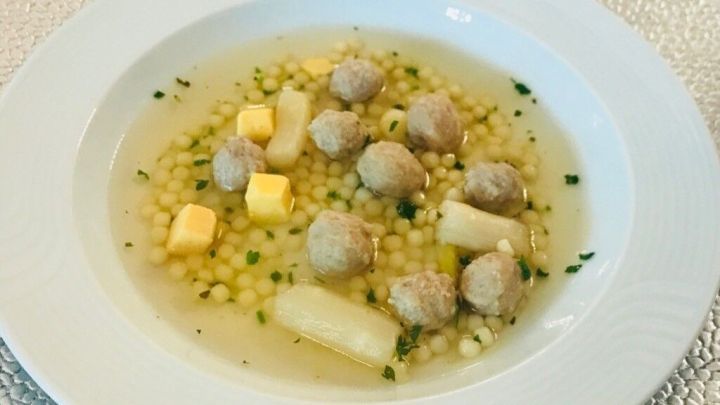 Kalbsleberkl&ouml;&szlig;chen schwimmen in einer klaren Br&uuml;he, umgeben von Gem&uuml;se und Kr&auml;utern auf einem eleganten Teller.