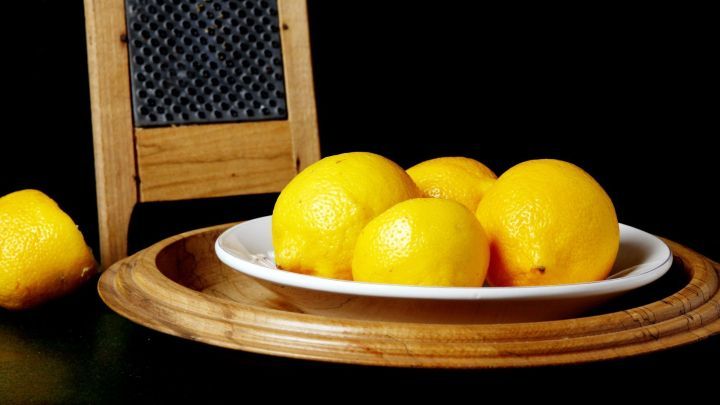 Eine Gruppe frischer Zitronen liegt auf einem wei&szlig;en Teller, umgeben von einem Holzbrett und einem Reibe-Geschirr, auf schwarzem Hintergrund.