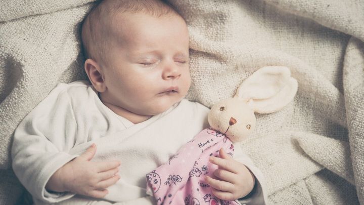 Schlafendes Baby h&auml;lt Schnuffeltuch mit Kuscheltierhase in pastellfarbenem Bett, Text auf Tuch: "Have a happy".