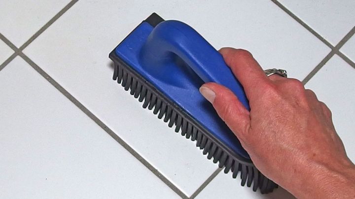 Eine Hand h&auml;lt eine blaue Reinigungsb&uuml;rste, w&auml;hrend sie &uuml;ber geflieste B&ouml;den im Badezimmer reinigt.