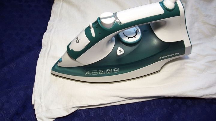 Ein Dampfb&uuml;geleisen b&uuml;geln gerade ein wei&szlig;es T-Shirt auf einem blauen Untergrund f&uuml;r ein schnelles Ergebnis.