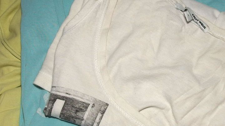 Ein gefaltetes wei&szlig;es T-Shirt liegt ordentlich auf einem blauen Shirt, umgeben von einer bunten W&auml;scheumgebung.