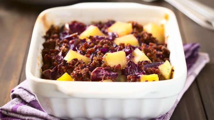 Ein Auflauf aus Hackfleisch, Rotkohl und Kartoffeln in einer wei&szlig;en Sch&uuml;ssel, auf einem lila Tuch auf einem Holztisch.