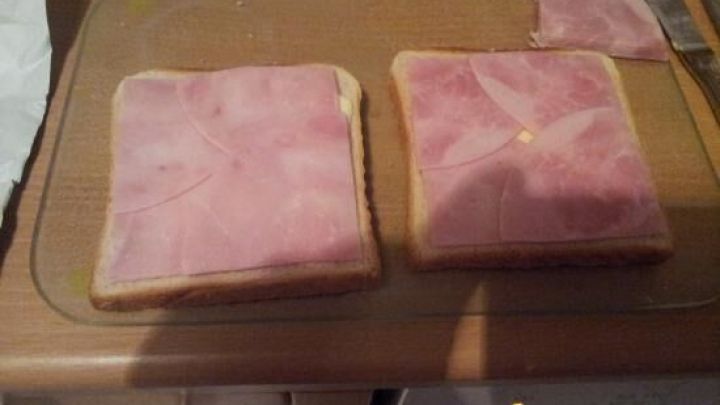 Runde Schinkenscheiben liegen auf eckigem Toast, der auf einem Holzbrett vorbereitet wird.