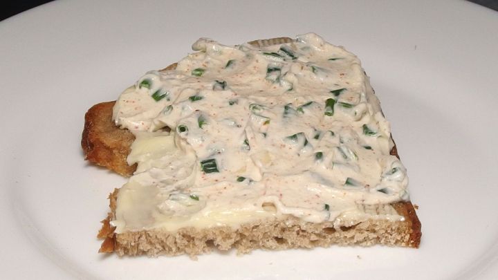 Ein St&uuml;ck Brot ist mit selbstgemachtem Fr&uuml;hlingsquark bedeckt, der mit Kr&auml;utern verfeinert ist, auf einem wei&szlig;en Teller.