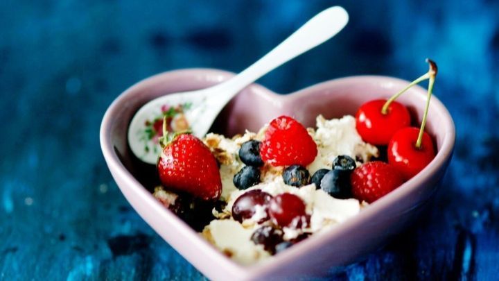 Eine herzf&ouml;rmige Schale mit cremigem Joghurt wird mit frischen Beeren und Kirschen dekoriert, auf einem blauen Hintergrund.