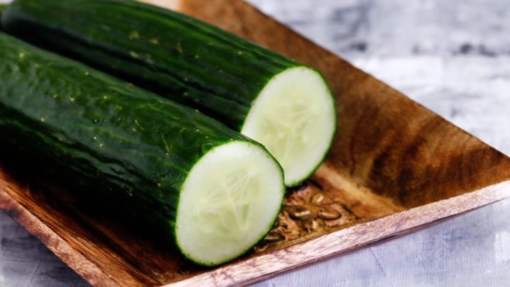 Zwei aufgeschnittene Schlangengurken liegen auf einem Holzbrett, umgeben von einer neutralen, grauen Oberfl&auml;che.