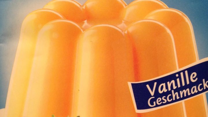 Ein Vanillepudding steht in einer Form, umgeben von frischen Erdbeeren, pr&auml;sentiert auf einer blauen Verpackung.