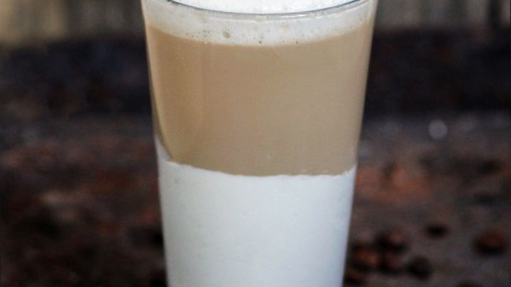 Ein Glas mit schaumiger Milch ist gef&uuml;llt, die obere Schicht ist hellbraun und die untere wei&szlig;, umgeben von Kaffeebohnen.