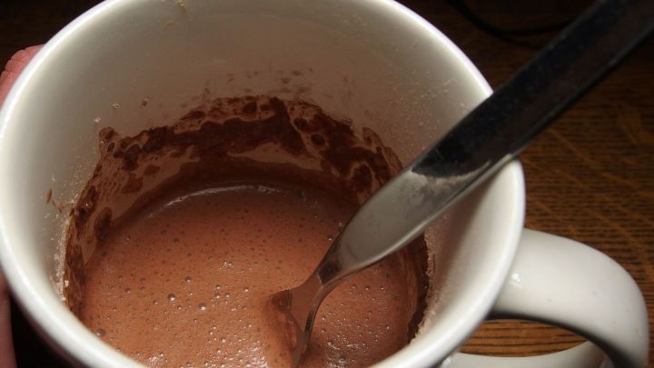 Ein gef&uuml;llter Becher mit Schokopudding, in dem ein L&ouml;ffel umr&uuml;hrt, steht auf einem Tisch in einer gem&uuml;tlichen Umgebung.