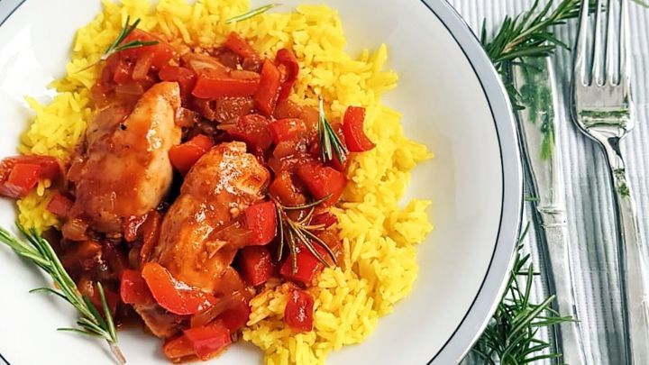 Ein Teller mit H&auml;hnchen in einem bunten Curry, serviert auf gelben Reis, umgeben von frischen Kr&auml;utern.