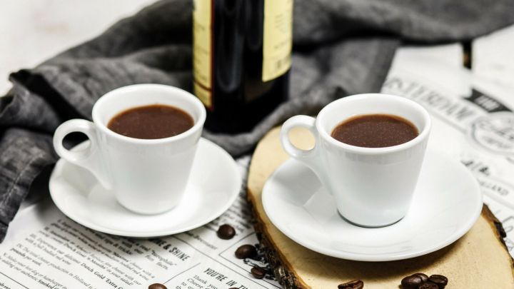 Zwei Tassen Kaffee stehen auf einem Holzbrett, umgeben von Kaffeebohnen und einer Weinflasche, auf einem Zeitungsblatt.