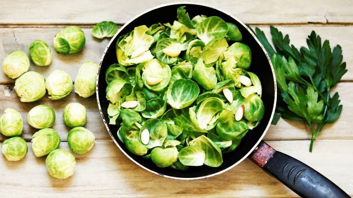 Gr&uuml;ne Rosenkohlbl&auml;tter liegen in einer Pfanne, umgeben von frischem Gem&uuml;se und Petersilie auf einer Holzoberfl&auml;che.