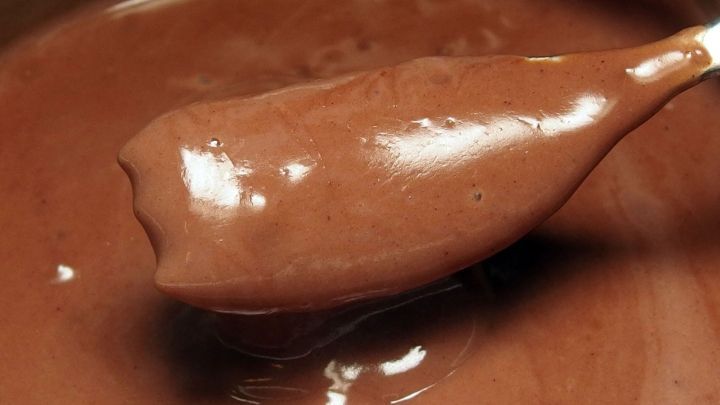 Eine gl&auml;nzende Schokoladenso&szlig;e wird auf einem L&ouml;ffel gehalten, w&auml;hrend sie in einer metallischen Sch&uuml;ssel ruht.