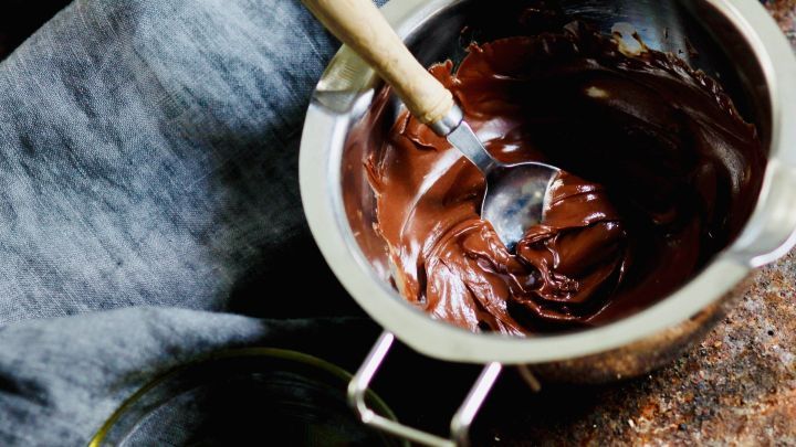 Ein silberner Topf mit geschmolzener Kuvert&uuml;re, darin r&uuml;hrt ein L&ouml;ffel; daneben steht ein Glas mit &Ouml;l auf einem Holztisch.