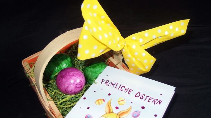 Ein Korb mit bunten Ostereiern und einem gelben Schleifenband steht auf einem Tisch, um den Ostertisch festlich zu dekorieren. Der Korb enth&auml;lt auch eine Karte mit dem Text "Frohe Ostern".