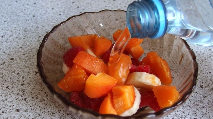 Frische Obstsalatst&uuml;cke werden mit sprudelndem Selterswasser in einer rustikalen Sch&uuml;ssel &uuml;bergossen, auf einer Tischoberfl&auml;che.