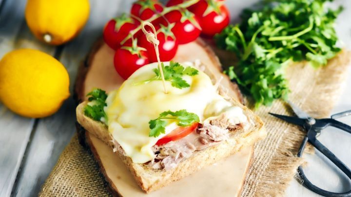 Ein belegter Toast mit K&auml;se, Tomate und Koriander liegt auf einem Holzbrett, umgeben von Zitronen und frischen Kr&auml;utern.
