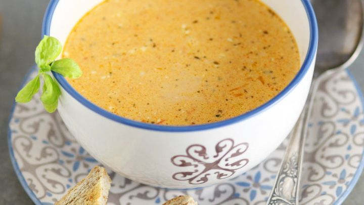 Ein cremiges Paprikas&uuml;ppchen f&uuml;llt eine Schale, umgeben von ger&ouml;stetem Brot und einer Zwiebel im Hintergrund.
