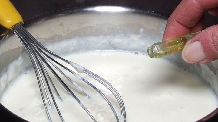 Ein Handr&uuml;hrger&auml;t vermischt cremigen Milchreis in einem Topf, w&auml;hrend eine Person einen gelben Kapselinhalt hinzuf&uuml;gt.