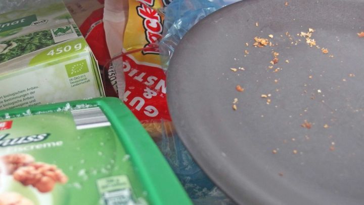 Auf einem grauen Teller sind Essensreste zu sehen, umgeben von Verpackungen, was Schimmel auf dem Geschirr f&ouml;rdern k&ouml;nnte.