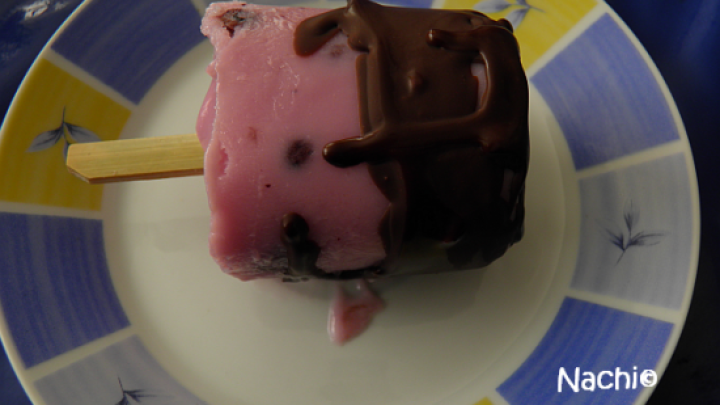 Ein gefrorener Joghurt am Stiel, halb in Schokolade getaucht, liegt auf einem bunten Teller. Text: "Nachie".