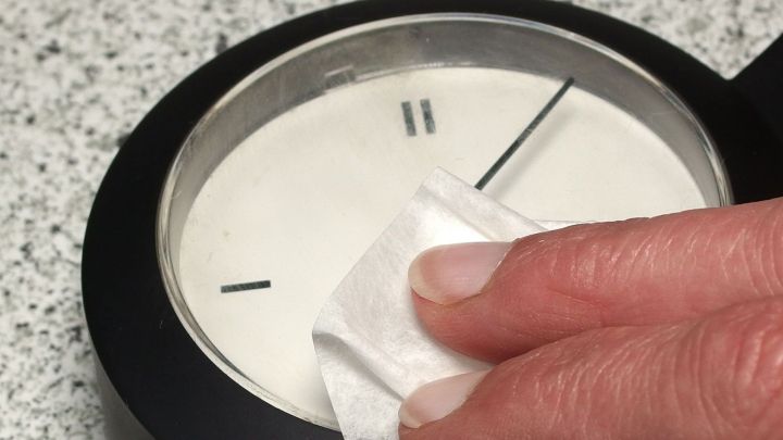 Ein Brillenputzt&uuml;chlein reinigt sorgf&auml;ltig das Ziffernblatt einer schwarz umrandeten Uhr auf einer wei&szlig;en Unterlage.