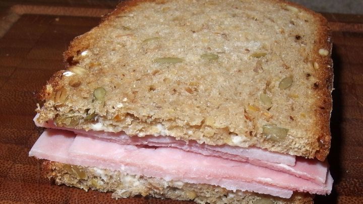 Ein Sandwich aus Vollkornbrot mit Schinken liegt auf einem Holzbrett, w&auml;hrend der Fokus auf der Reduzierung von Schulbrotverschwendung liegt.