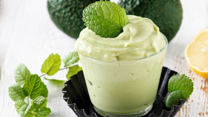 Eine cremige Avocado-Nachspeise steht in einem Glas, umgeben von frischen Minzbl&auml;ttern und einer halben Zitrone.