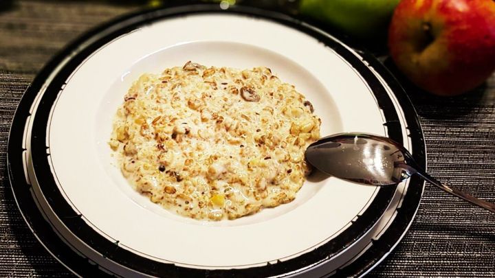 Ein M&uuml;sli-Brei auf einem wei&szlig;en Teller mit einem L&ouml;ffel. Im Hintergrund sind ein gr&uuml;ner Apfel und ein roter Apfel sichtbar.
