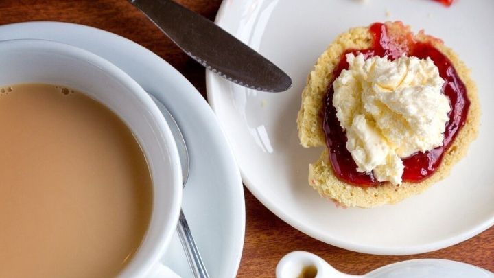 Eine Tasse Kaffee steht neben einem Teller mit Scones, die mit Marmelade und clotted cream belegt sind, auf einem Holztisch.