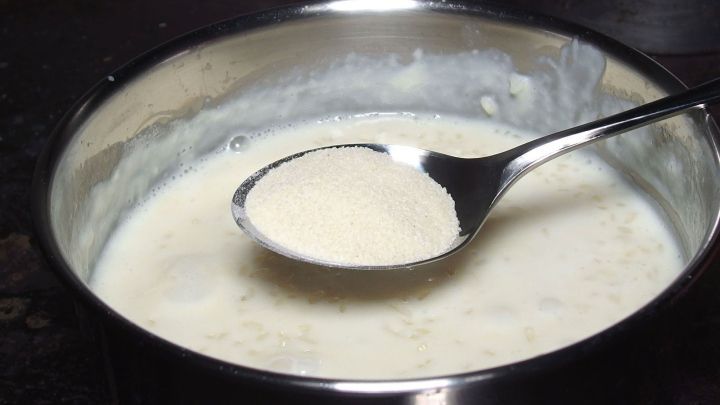 Ein Topf mit cremigem Milchreis steht auf einem Tisch, w&auml;hrend ein L&ouml;ffel darin r&uuml;hrt und die feine Konsistenz zeigt.