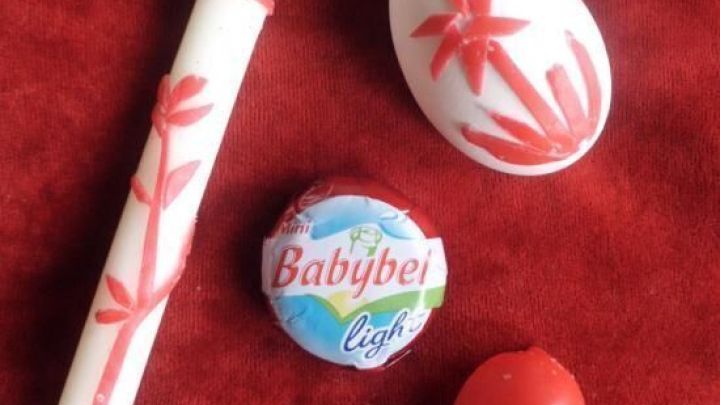 Ein roter Tisch mit verschiedenen (Oster)-Basteleien aus Babybel-Wachs, darunter eine dekorierte Kerze und ein Ei.