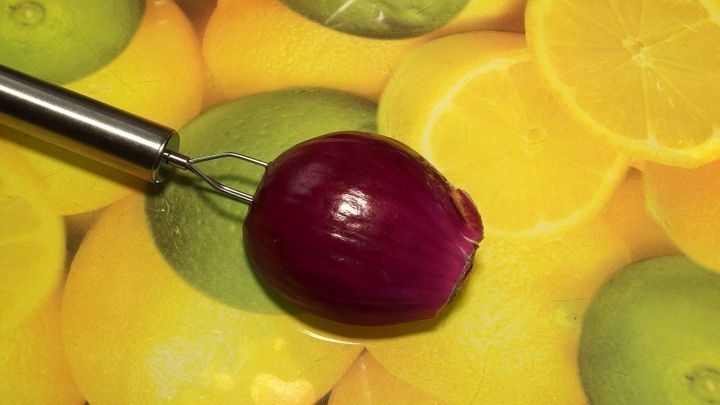 Eine rote Zwiebel liegt auf einem bunten Tuch mit Zitronen, w&auml;hrend ein K&uuml;chenwerkzeug sie zum Schneiden h&auml;lt.